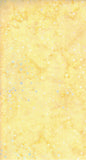 A close-up of a pale yellow cotton batik fabric with subtle marbling and scattered small light blue and white speckles.