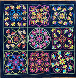 A square dark blue quilt featuring a 3x3 grid of colorful floral and geometric applique motifs in pink, yellow, orange, green, and blue, photographed flat against a wood floor background.